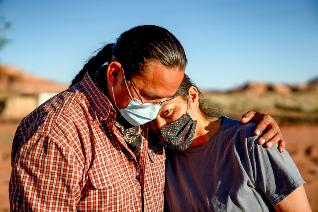 A man and woman wearing a mask and embracing in front of an open blue sky.